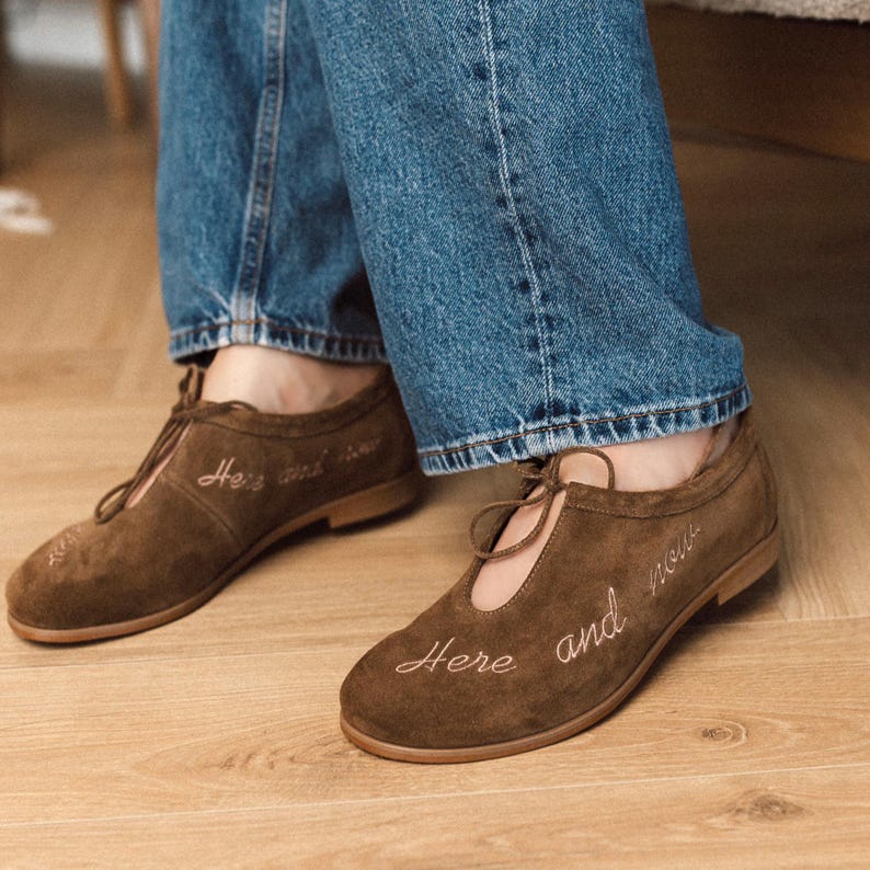 Pu&ograve; includere: Scarpe in camoscio marrone con tacco basso e design stringato. Le parole "Here and now" sono ricamate sulle scarpe. Le scarpe sono indossate con jeans blu.