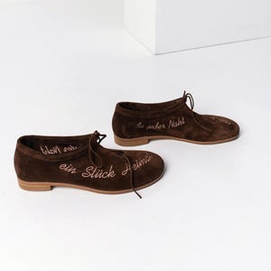 Pu&ograve; includere: Un paio di scarpe in camoscio marrone con lacci. Le scarpe hanno suole marrone chiaro e presentano un testo ricamato in un colore marrone pi&ugrave; chiaro. Il testo sulle scarpe recita "ein St&uuml;ck Heimat" e "In jeder Naht".