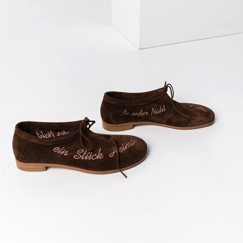 Pu&ograve; includere: Un paio di scarpe in camoscio marrone con lacci. Le scarpe hanno suole marrone chiaro e presentano un testo ricamato in un colore marrone pi&ugrave; chiaro. Il testo sulle scarpe recita "ein St&uuml;ck Heimat" e "In jeder Naht".