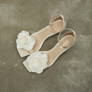 May include: A pair of white heeled sandals with a large white flower embellishment on each strap. The sandals have an ankle strap with the brand name "LOOLIE" written on it.