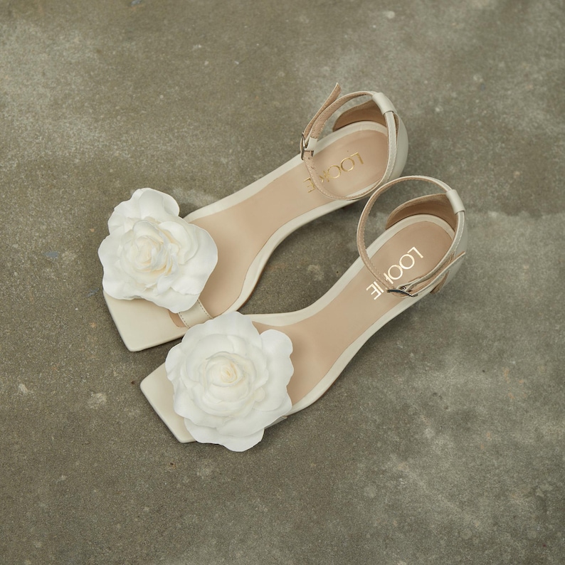 May include: A pair of white heeled sandals with a large white flower embellishment on each strap. The sandals have an ankle strap with the brand name "LOOLIE" written on it.