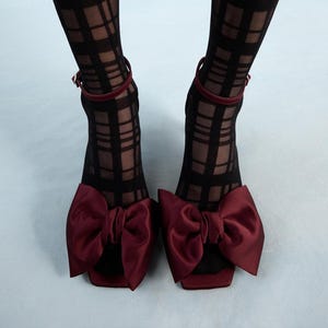 Op de afbeelding: Een paar bordeauxkleurige sandalen met hoge hakken en grote satijnen strikken. De sandalen worden gedragen met zwart-wit geblokte panty's.