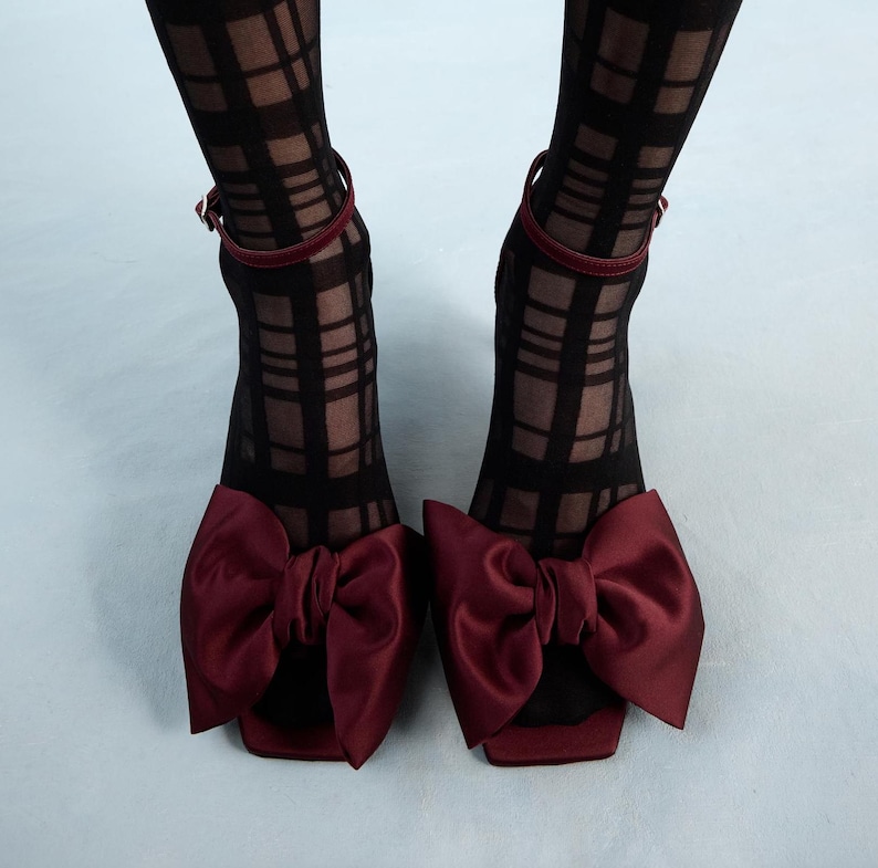 Op de afbeelding: Een paar bordeauxkleurige sandalen met hoge hakken en grote satijnen strikken. De sandalen worden gedragen met zwart-wit geblokte panty's.