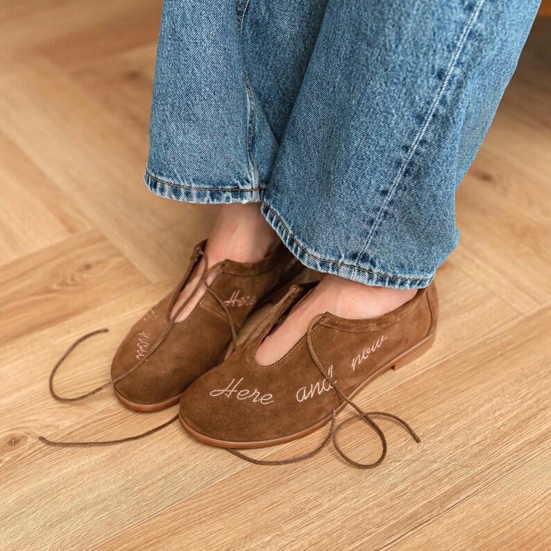 Pu&ograve; includere: Scarpe in camoscio marrone con un design scollato e lacci lunghi. Le scarpe hanno le parole "Here and now" ricamate in corsivo sulla parte superiore. Indossate con jeans blu.
