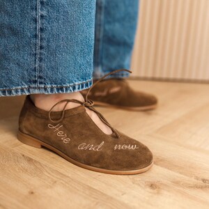 Pu&ograve; includere: Scarpe in camoscio marrone con tacco basso e design stringato. Le parole "Here and now" sono ricamate in bianco sul lato della scarpa. Le scarpe sono indossate con jeans blu.