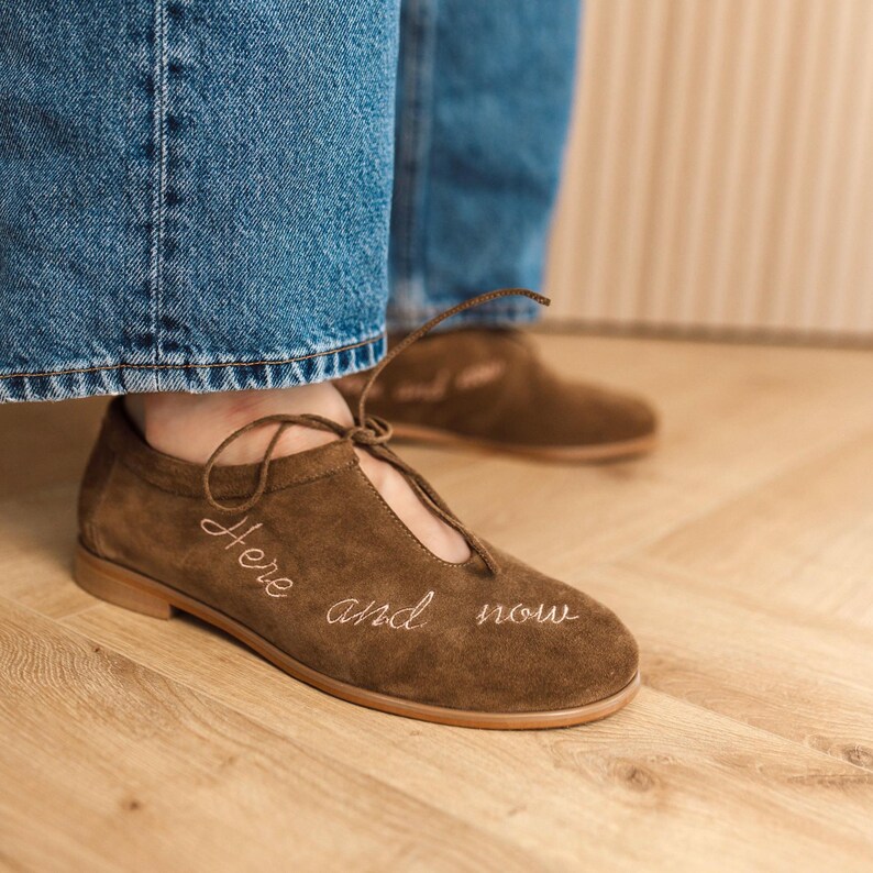 Pu&ograve; includere: Scarpe in camoscio marrone con tacco basso e design stringato. Le parole "Here and now" sono ricamate in bianco sul lato della scarpa. Le scarpe sono indossate con jeans blu.