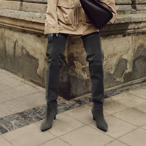 May include: A person wearing a tan jacket and dark gray suede over-the-knee boots. The boots have a pointed toe and a low heel.