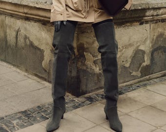Könnte beinhalten: Eine Person trägt eine braune Jacke und dunkelgraue Overknee-Stiefel aus Wildleder. Die Stiefel haben eine spitze Zehenpartie und einen niedrigen Absatz.