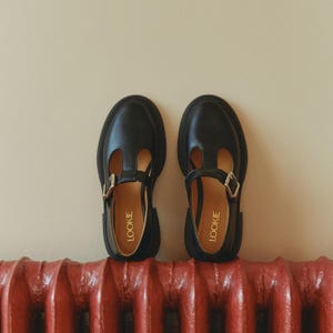 May include: A pair of black leather shoes with a buckle and a cut-out design. The shoes are sitting on top of a red radiator. The brand name "LOOKIE" is printed on the inside of each shoe.