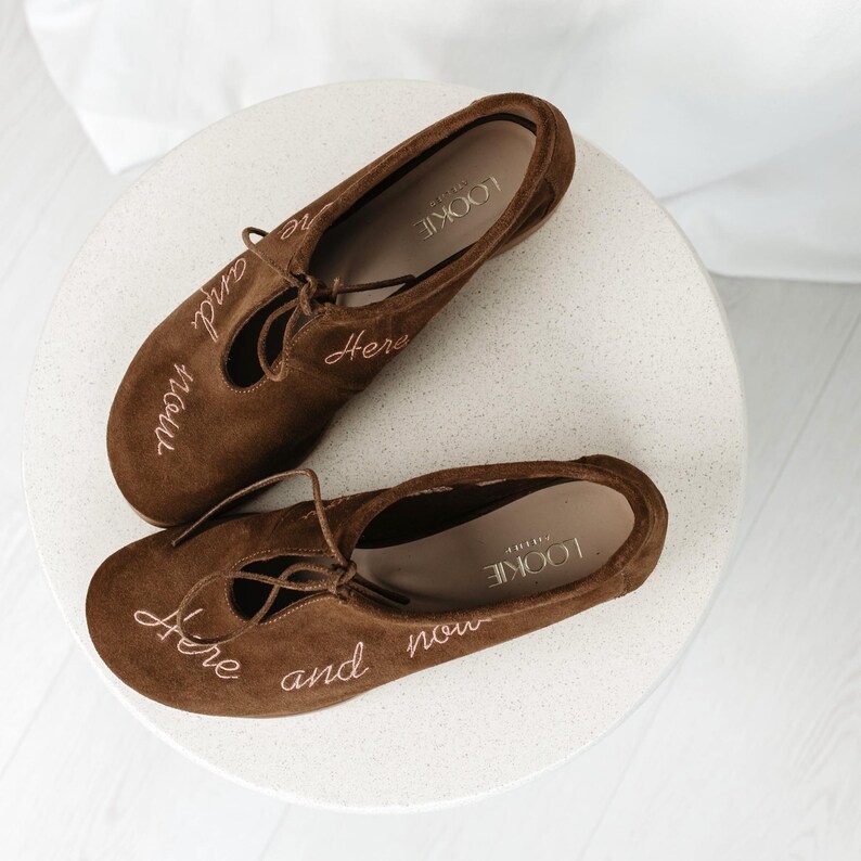 Pu&ograve; includere: Ballerine in camoscio marrone con design stringato. Le scarpe hanno le parole "Here and now" ricamate in un marrone pi&ugrave; chiaro. Il marchio "Lookie" &egrave; visibile all'interno della scarpa.