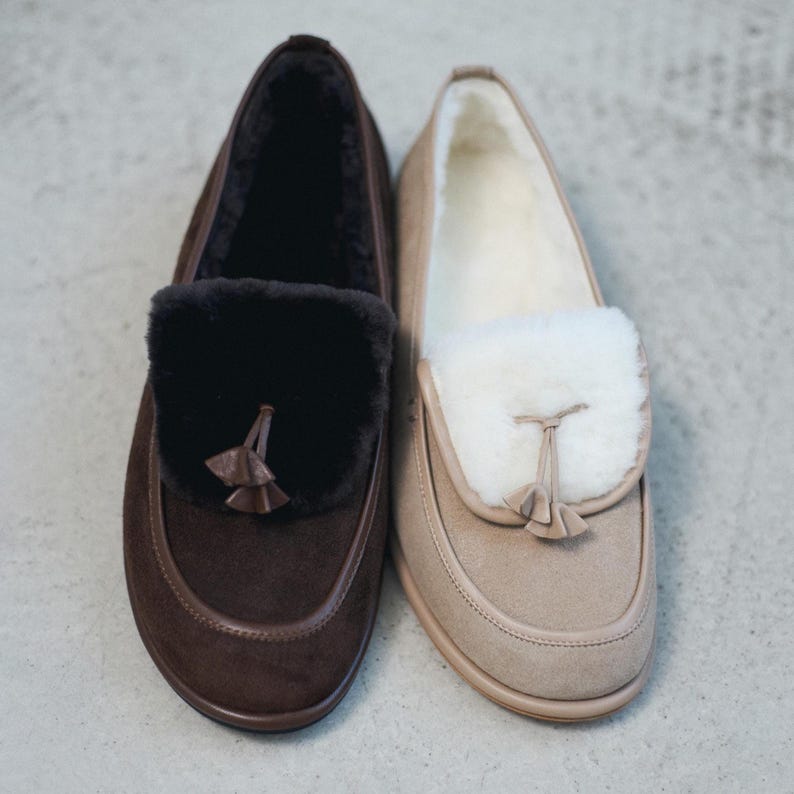 May include: Two pairs of suede loafers. One is dark brown with a black fur accent and tassel. The other is tan with a cream fur accent and tassel. Both have a rounded toe and a soft, comfortable appearance.