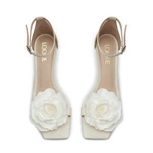 May include: A pair of white heeled sandals with a large white flower embellishment on the toe. The sandals have an ankle strap with the word "LOOKIE" written on it.
