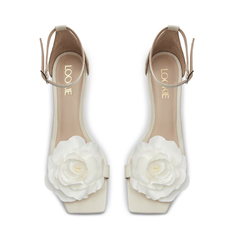 May include: A pair of white heeled sandals with a large white flower embellishment on the toe. The sandals have an ankle strap with the word "LOOKIE" written on it.