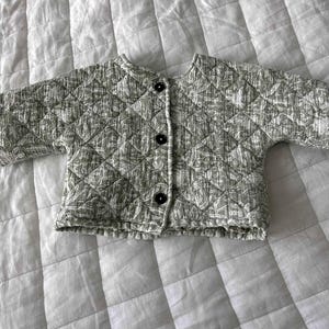 May include: A quilted jacket in a light green and white floral pattern. The jacket has a round collar, three dark buttons, and short sleeves. The jacket is made of a soft, textured fabric.