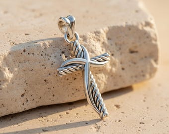 May include: A silver-colored cross pendant with a twisted rope design. The cross is displayed against a light-colored, textured stone surface. The pendant has a heart-shaped bail for a chain.