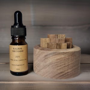 May include: A brown glass bottle with a black dropper top and a label that reads "Ralph's Orchard Zen Cassis, Rose, Green Tea Luxury Fragrance Oil 10ml". The bottle is next to a wooden diffuser with wooden sticks.