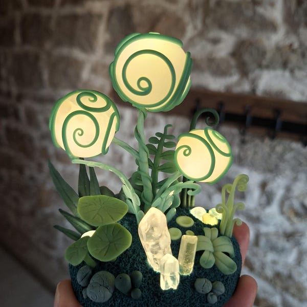 Könnte beinhalten: Eine leuchtende Fantasy-Skulptur mit drei leuchtenden, kugelförmigen Blumenformen mit grünen Spiralakzenten. Die Skulptur umfasst grüne Blätter, Stiele und Kristalle, alles auf einem dunkelblauen Sockel. Das Design erinnert an ein Märchen.