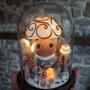 Op de afbeelding: Een grillige paddenstoelvormige sculptuur in een glazen stolp. De sculptuur heeft een bruine paddenstoelhoed met wervelende patronen, een rond gezicht en een blauw outfit. De scène bevat miniatuur paddenstoelen, bladeren en lichtjes.
