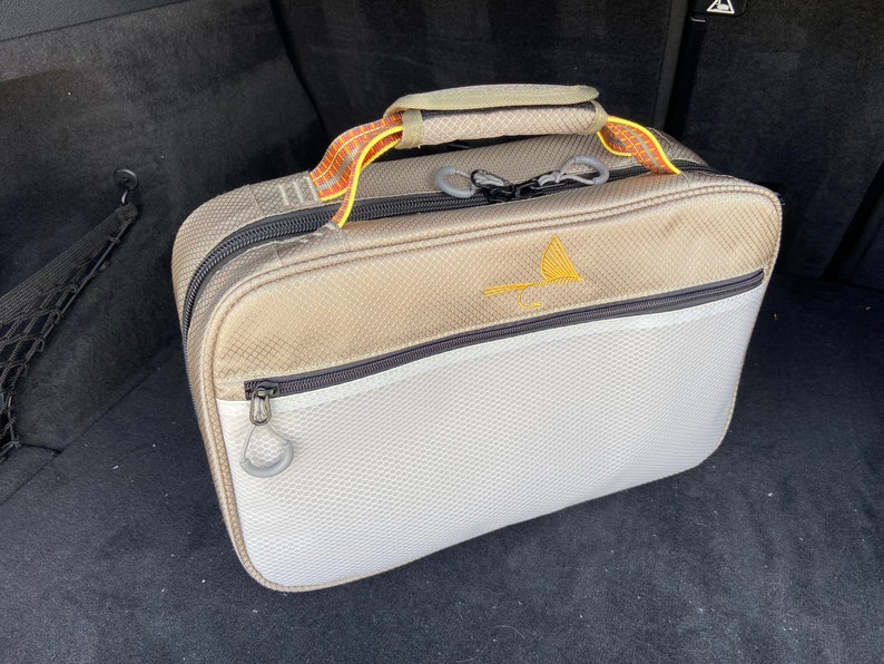 May include: A gray and white travel bag with a black zipper and a yellow and orange handle. The bag has a mesh pocket on the front and a logo on the top.