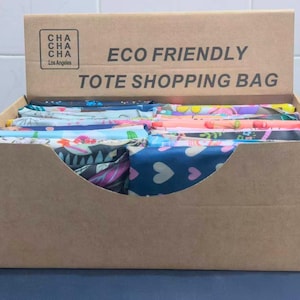 May include: A cardboard display box filled with folded tote shopping bags. The box has the text "ECO FRIENDLY TOTE SHOPPING BAG" printed on the front. The bags have various colorful floral and heart patterns.