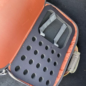 May include: Open black carrying case with a brown interior and a gray foam insert with multiple circular cutouts. The case has a zipper closure and a gray strap.