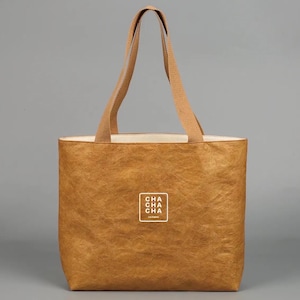 May include: A brown tote bag with beige handles and a white interior. The bag features the text "CHA CHA CHA" in a white square on the front. The bag is made of a textured material and is designed for carrying items.