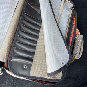 May include: A gray and brown zippered carrying case with a black zippered pocket and a white mesh pocket. The case has a black strap with a snap closure.