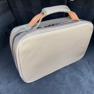 May include: A beige travel bag with a black zipper and orange and brown straps. The bag is sitting in the back of a car.