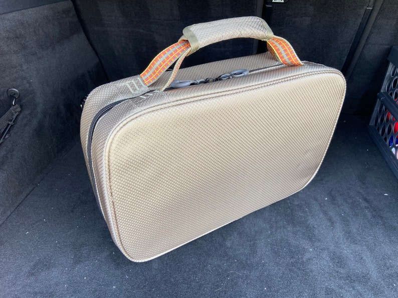 May include: A beige travel bag with a black zipper and orange and brown straps. The bag is sitting in the back of a car.