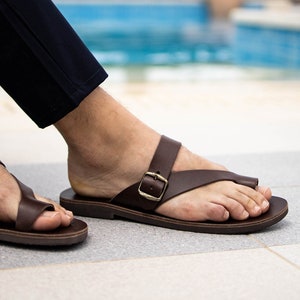 May include: A pair of brown leather sandals with a buckle strap. The sandals are worn by a person sitting by a pool.