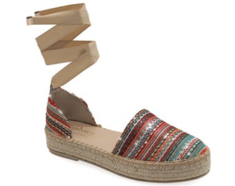 May include: Espadrille sandal with a multi-colored striped fabric upper, featuring red, orange, and teal hues. The shoe has a woven jute sole and a beige ribbon ankle tie. The sandal is designed for a casual, warm-weather look.