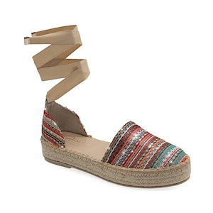 May include: Espadrille sandal with a multi-colored striped fabric upper, featuring red, orange, and teal hues. The shoe has a woven jute sole and a beige ribbon ankle tie. The sandal is designed for a casual, warm-weather look.