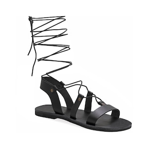 May include: Black leather sandals with lace-up ties that wrap around the ankle. The sandals have a flat sole and a simple design.