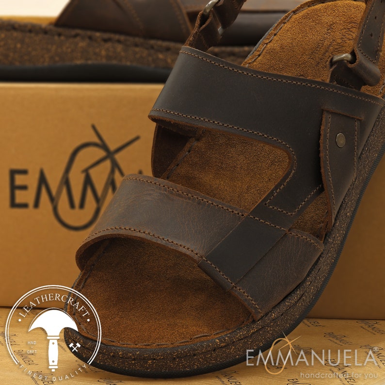 May include: Brown leather sandals with multiple straps and a cork footbed. The sandals have adjustable buckles and are designed for comfort. The image also shows the brand name "Emmanuela" and the words "Leathercraft" and "Finest Quality".