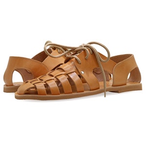 May include: A pair of light brown leather sandals with a woven design on the top. The sandals have a flat sole and are laced up with light brown laces. The open-toe sandals have cut-out details on the sides.