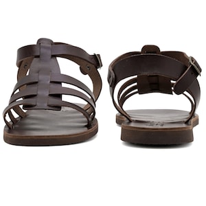 May include: Two brown leather sandals with multiple straps and buckles. The sandals have a flat sole and a classic gladiator style. The leather appears to be a rich, dark brown color, and the sandals are displayed against a white background.