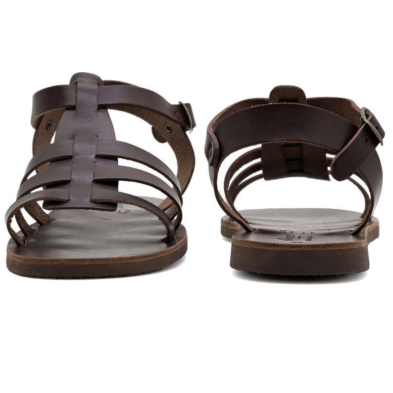 May include: Two brown leather sandals with multiple straps and buckles. The sandals have a flat sole and a classic gladiator style. The leather appears to be a rich, dark brown color, and the sandals are displayed against a white background.