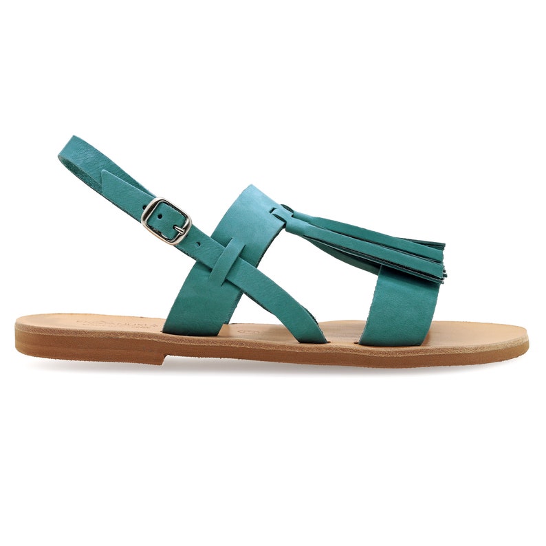 May include: Teal leather sandals with a flat sole. The sandals have a T-strap design, a buckle fastening, and a decorative tassel. The footbed is light brown.