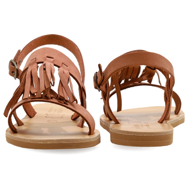 May include: A pair of brown leather sandals with fringe detailing. The sandals feature multiple straps and a buckle fastening. The soles are a light tan colour, and the sandals are set against a white background.