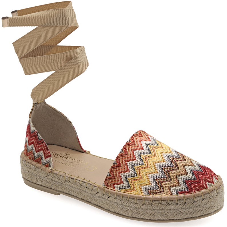May include: Espadrille sandal with a colorful chevron pattern in red, yellow, and beige. The shoe has a woven jute sole and a beige ankle wrap. The brand name "EMMANUELLE" is visible on the insole.