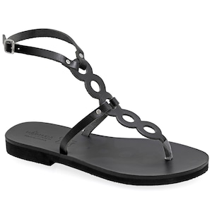 May include: Black leather sandals with a delicate ankle strap featuring a circle cut-out design. The sandals have a flat sole and are made in Greece.