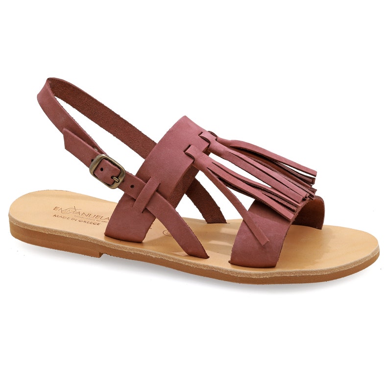 May include: Dusty rose leather sandals with a strap and buckle closure. The sandals feature a decorative fringe detail and a light brown sole. The words "EMMANUELA MADE IN GREECE" are printed on the sole.
