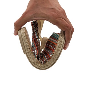 May include: A pair of flexible espadrille flats. The shoes have a woven jute sole and a colorful sequined upper with red, teal, and orange stripes. The shoes are being bent to show their flexibility.