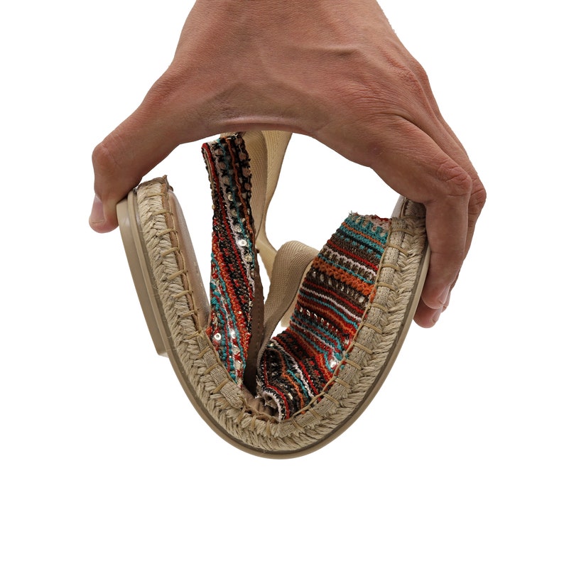 May include: A pair of flexible espadrille flats. The shoes have a woven jute sole and a colorful sequined upper with red, teal, and orange stripes. The shoes are being bent to show their flexibility.
