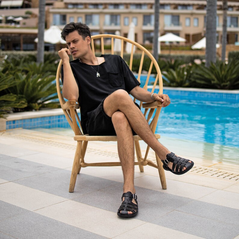 May include: Black leather sandals with multiple straps and buckles. The sandals are worn by a person sitting in a light-colored wooden chair. The person is wearing a black shirt and shorts. The background includes a swimming pool and palm trees.