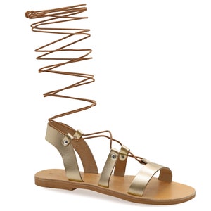 May include: Gold leather sandals with brown laces that tie up the leg. The sandals have a flat sole and a simple design.