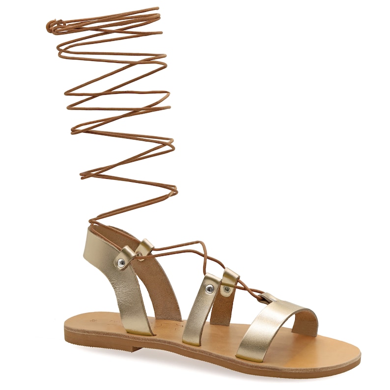 May include: Gold leather sandals with brown laces that tie up the leg. The sandals have a flat sole and a simple design.