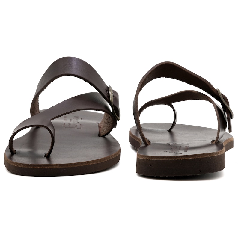 May include: Brown leather sandals with two straps and a buckle. The sandals have a toe loop and a flat sole.