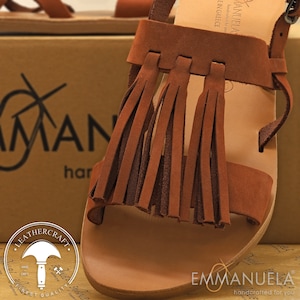 May include: Brown leather sandals with fringe detail. The sandals are handcrafted in Greece and feature a leather sole. The brand name "EMMANUELA" is printed on the sole.