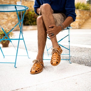May include: A pair of light brown leather sandals with a woven design. The sandals have tan laces and are worn by a person sitting on a blue chair. The sandals are a stylish footwear choice for warm weather.
